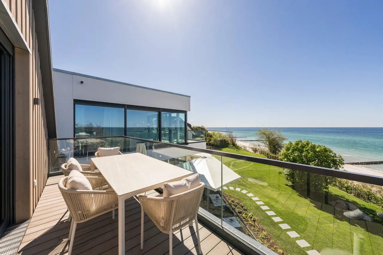 Sonniger Balkon mit Esstisch, Stühlen, Glasgeländer und Panoramablick auf das Meer.